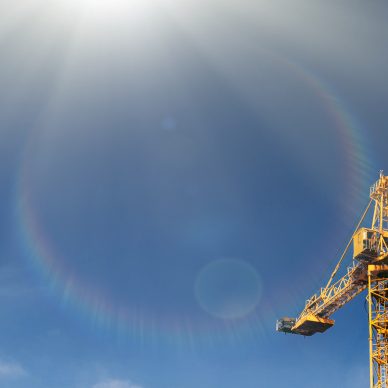 Construction crane tower in sun light beams on background of  blue sky. Crane and building working progress on construction site. Yellow lifting faucet.  Space for text